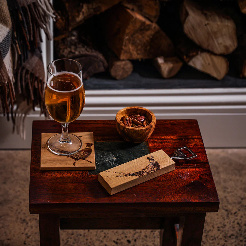 Glass of amber liquid on a wooden coaster with a bowl and bottle opener on a small table against a fireplace.