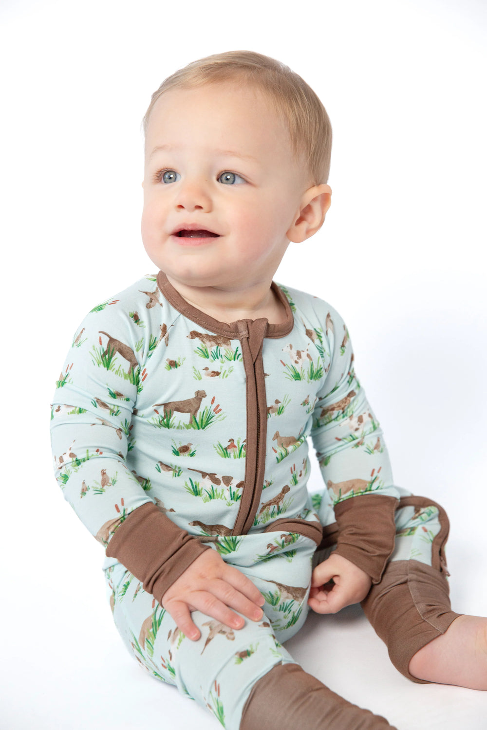 A baby sitting and wearing a long-sleeve footed sleeper with a light blue-green background, brown trim, and a woodland animal print, photographed against a white studio background.