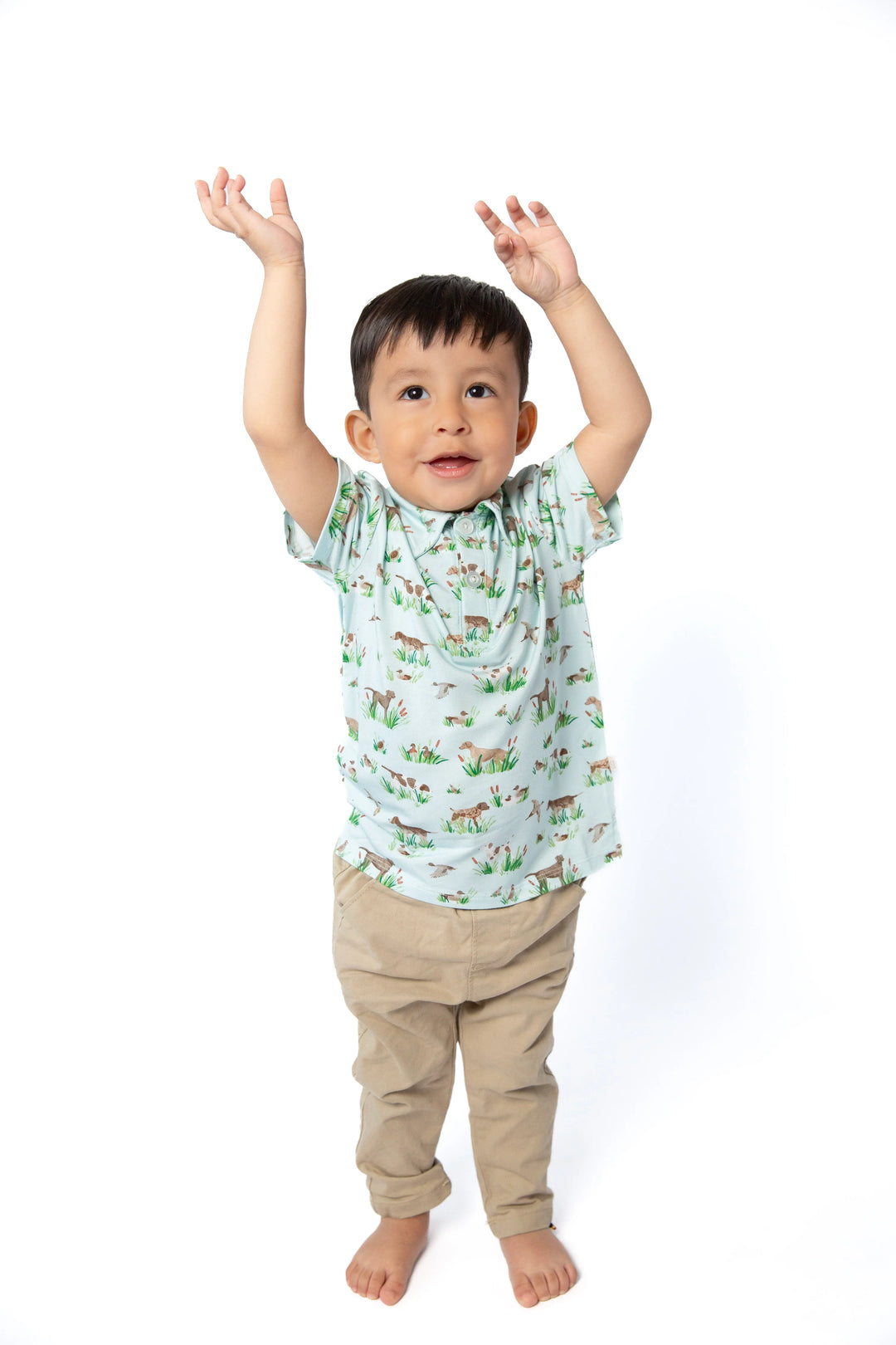 A toddler boy stands barefoot against a white background with his arms raised, wearing a light green short-sleeve t-shirt with a colorful animal print and beige pants.