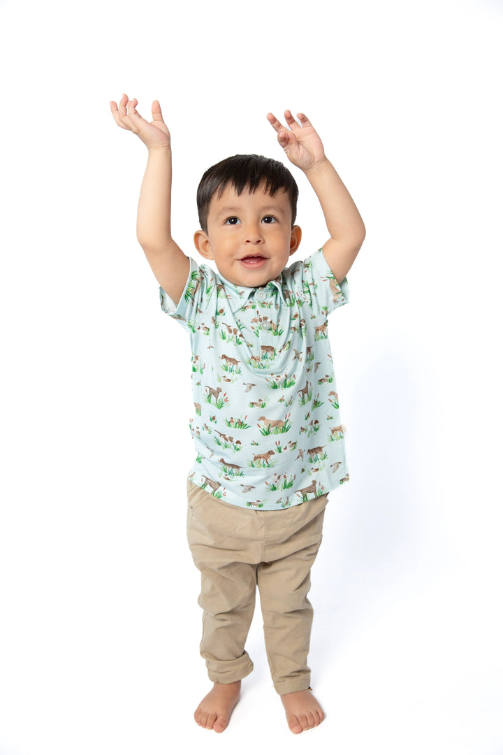 A toddler boy stands barefoot against a white background with his arms raised, wearing a light green short-sleeve t-shirt with a colorful animal print and beige pants.