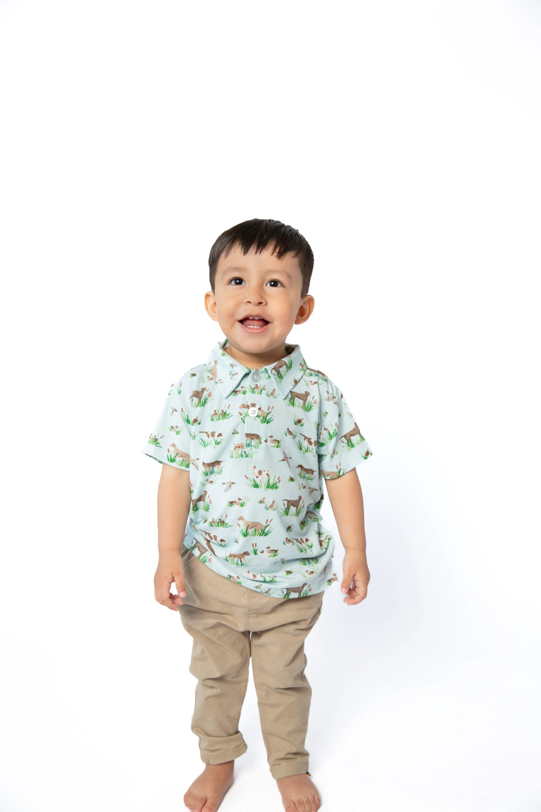 A toddler boy standing against a white background, wearing a light mint green short-sleeve button-up shirt with a colorful woodland animal print and khaki pants.