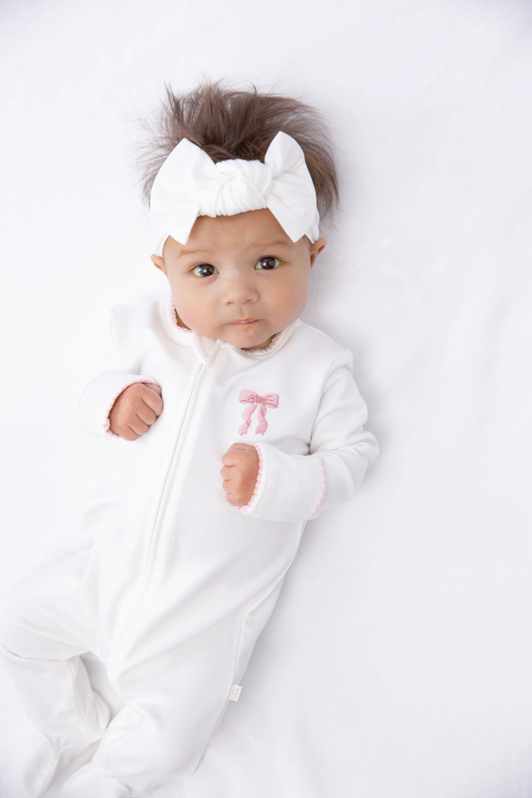 A baby lying on a white blanket, wearing a white footed onesie with a small pink bow on the chest and a matching white bow headband.