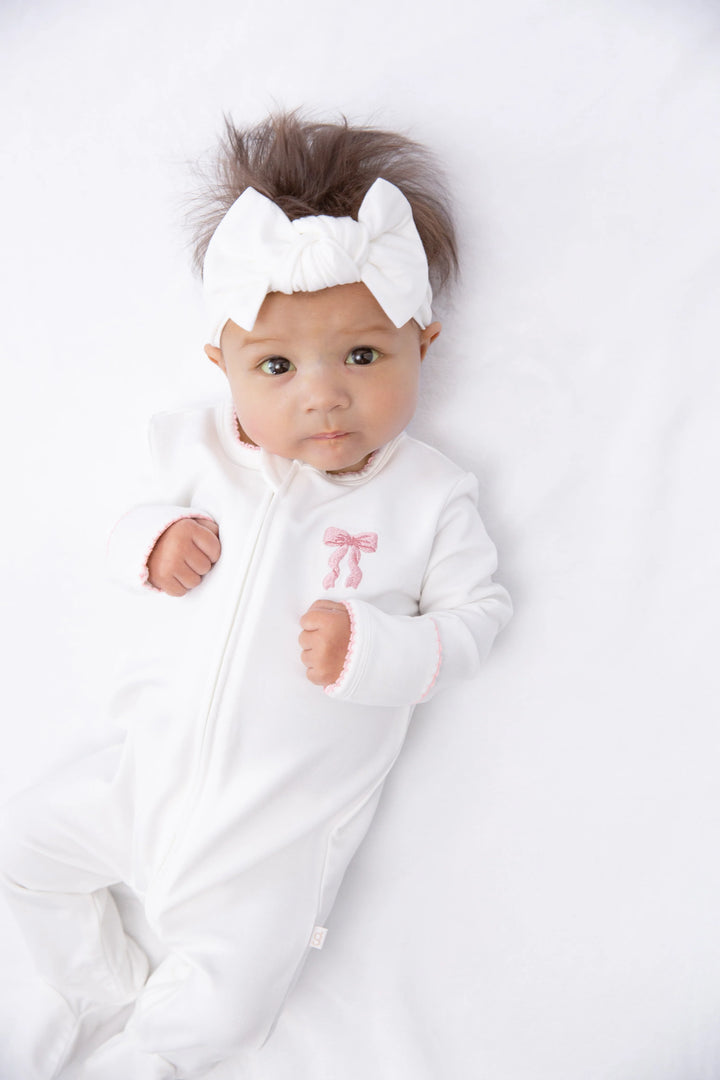 A baby lying on a white blanket, wearing a white footed onesie with a small pink bow on the chest and a matching white bow headband.