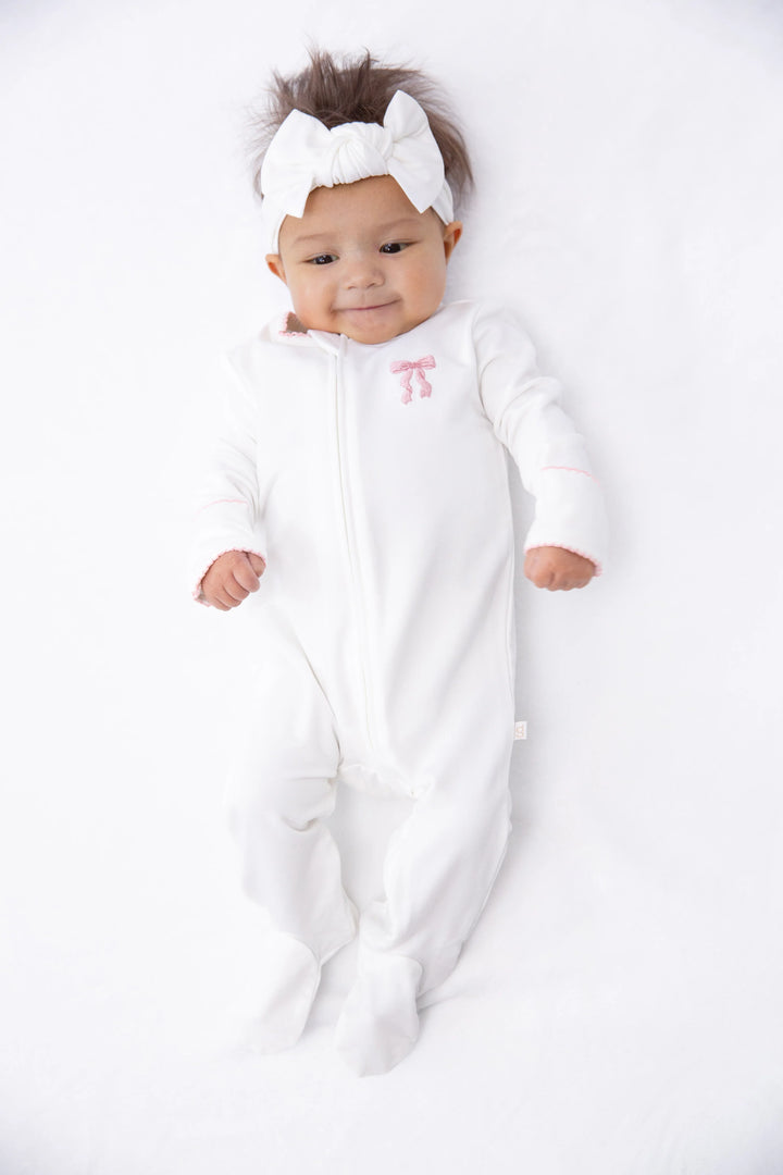 A baby lying on a white blanket wearing a white footed sleeper with a small pink bow detail on the chest, along with a matching white bow headband.