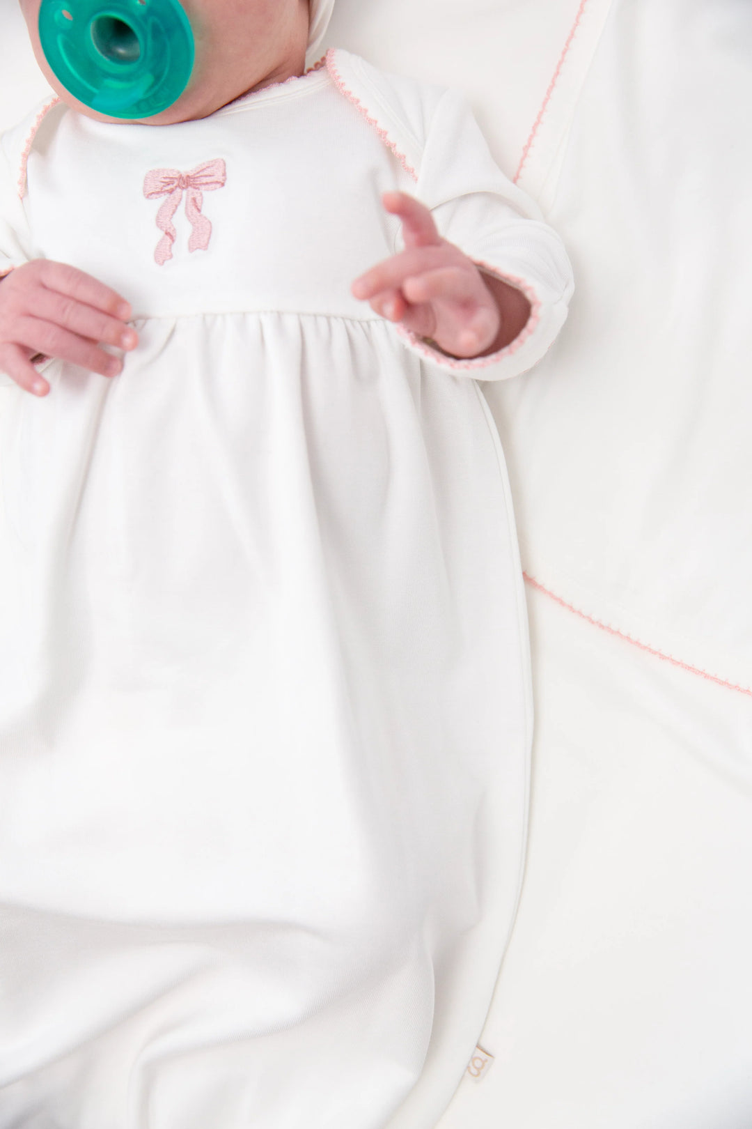 A close-up of a baby wearing a white long-sleeve swaddle or sleep sack with a small pink bow embroidery on the chest, lying on a white blanket. The baby's hands and one hand are visible, and a teal pacifier is in the mouth.