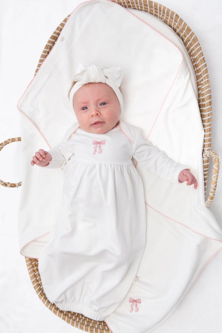 A baby lying on their back inside a woven bassinet, wearing a white long-sleeve dress with a small pink bow on the chest and a matching white bow headband.