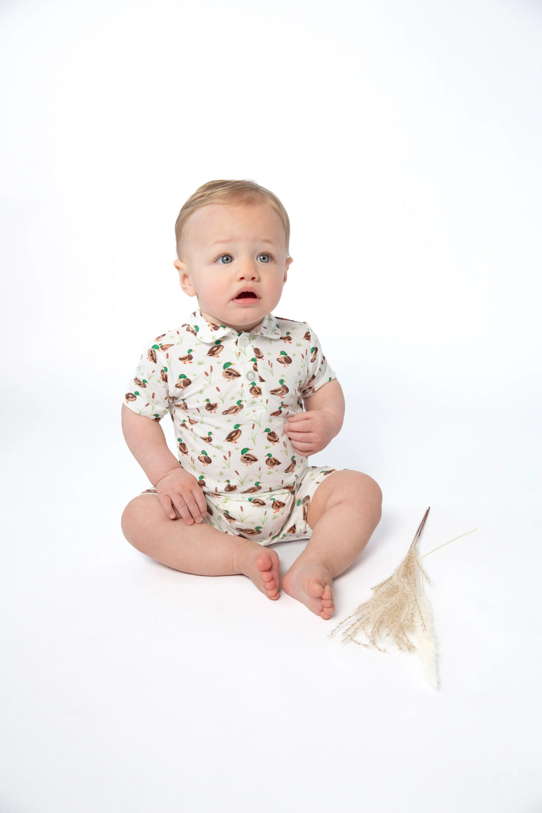 A baby sitting cross-legged on a white studio background, wearing a short-sleeve cream romper with a small multicolor floral print. A dried decorative plant stem lies beside the baby.