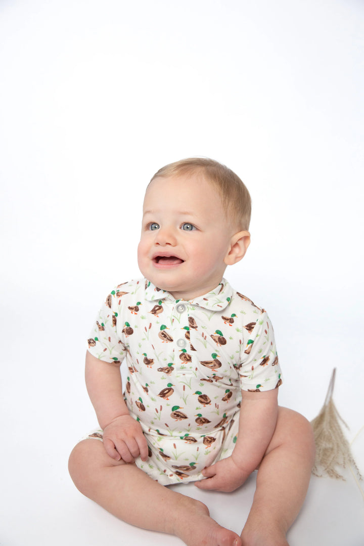 A baby sitting on a white studio background wearing a short-sleeve collared romper with a brown and green bird print on a cream base.