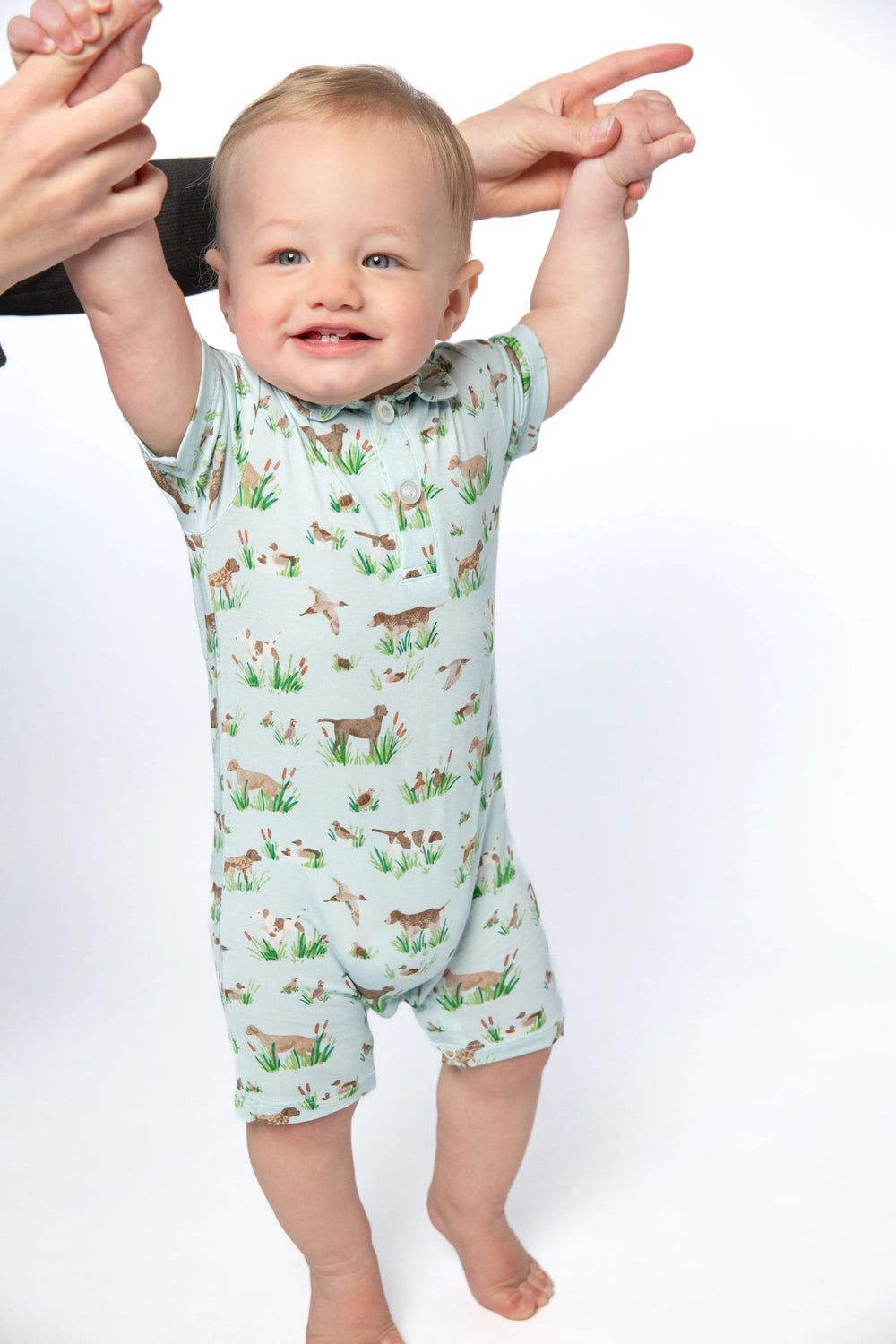 A baby wearing a short-sleeve light green romper with a woodland deer print, held up by an adult's hands against a white studio background.