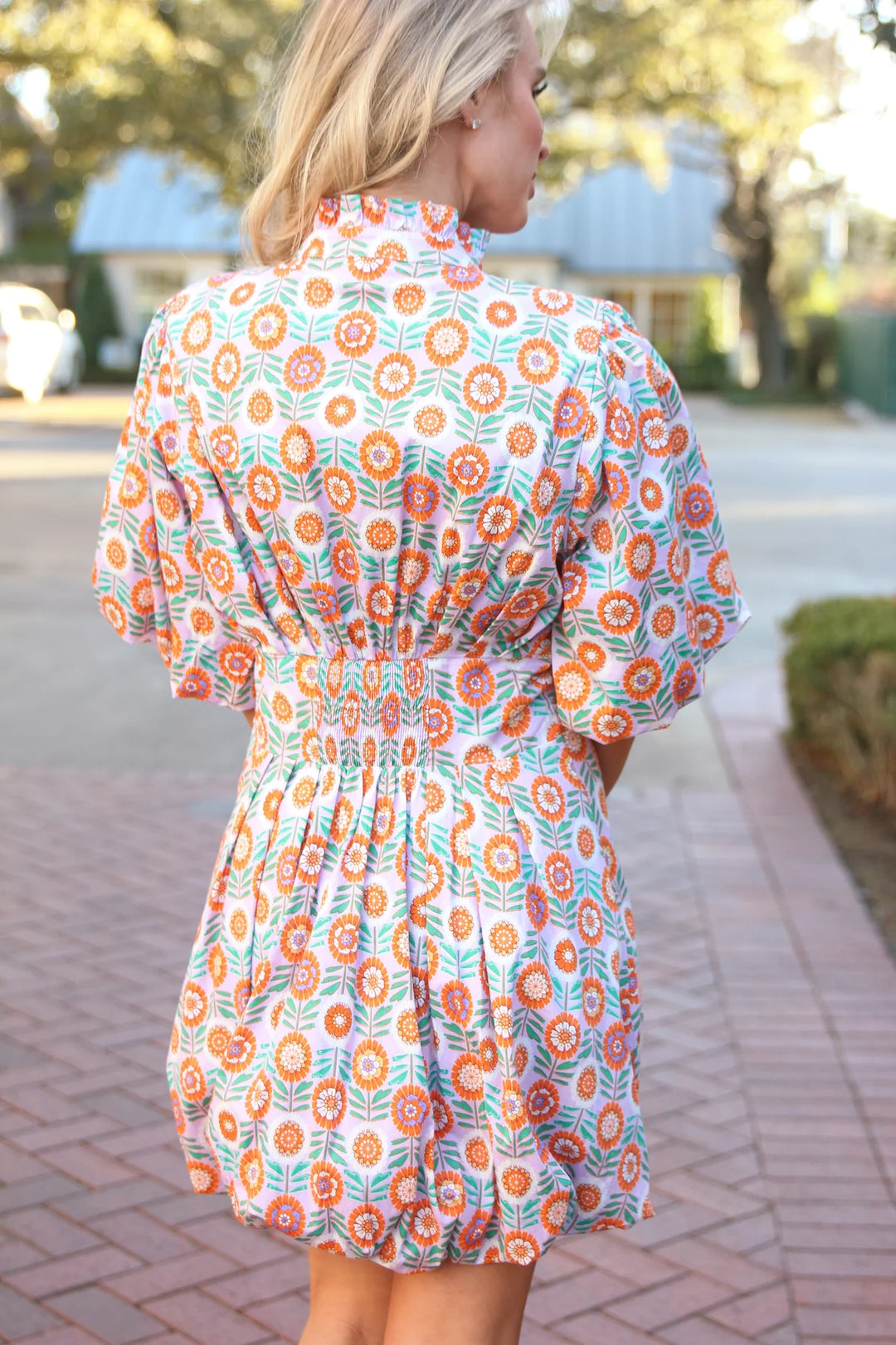 Woman wearing a floral dress walking on a sidewalk with trees and buildings in the background
