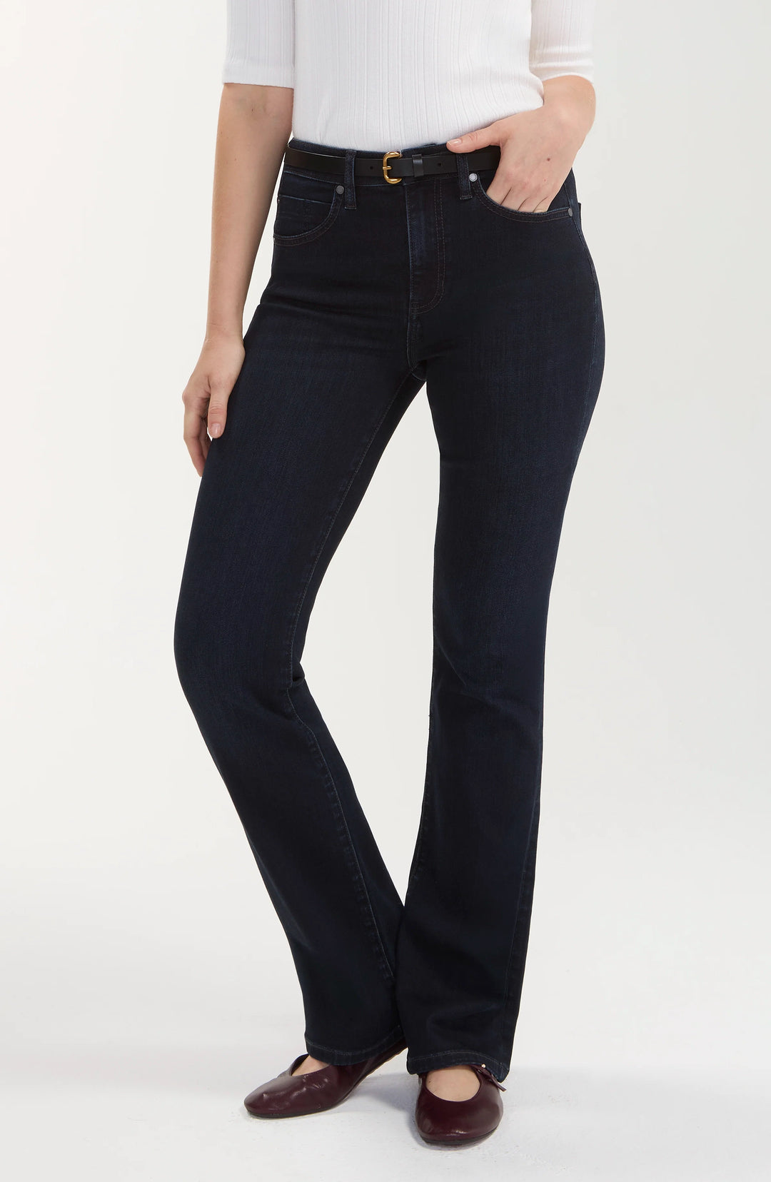 A model wearing a pair of dark wash flare jeans, styled with a white ribbed top and dark brown leather flats, photographed against a clean white studio background.