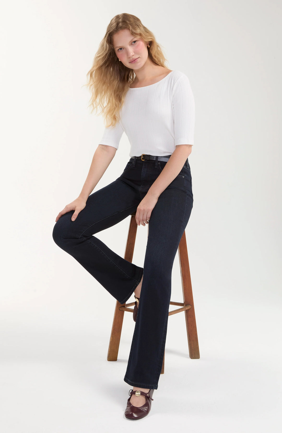 A woman models a white short-sleeve top and dark navy wide-leg jeans while sitting on a wooden stool against a plain white studio background.