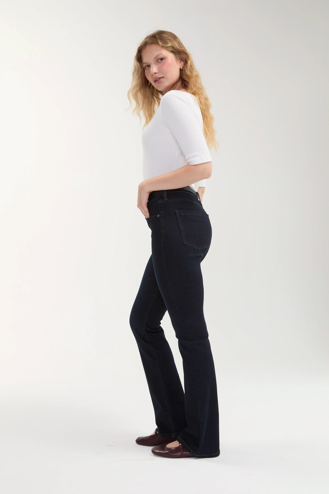 A woman modeling dark wash flared jeans with a white short-sleeve top, standing in profile against a light gray studio background.