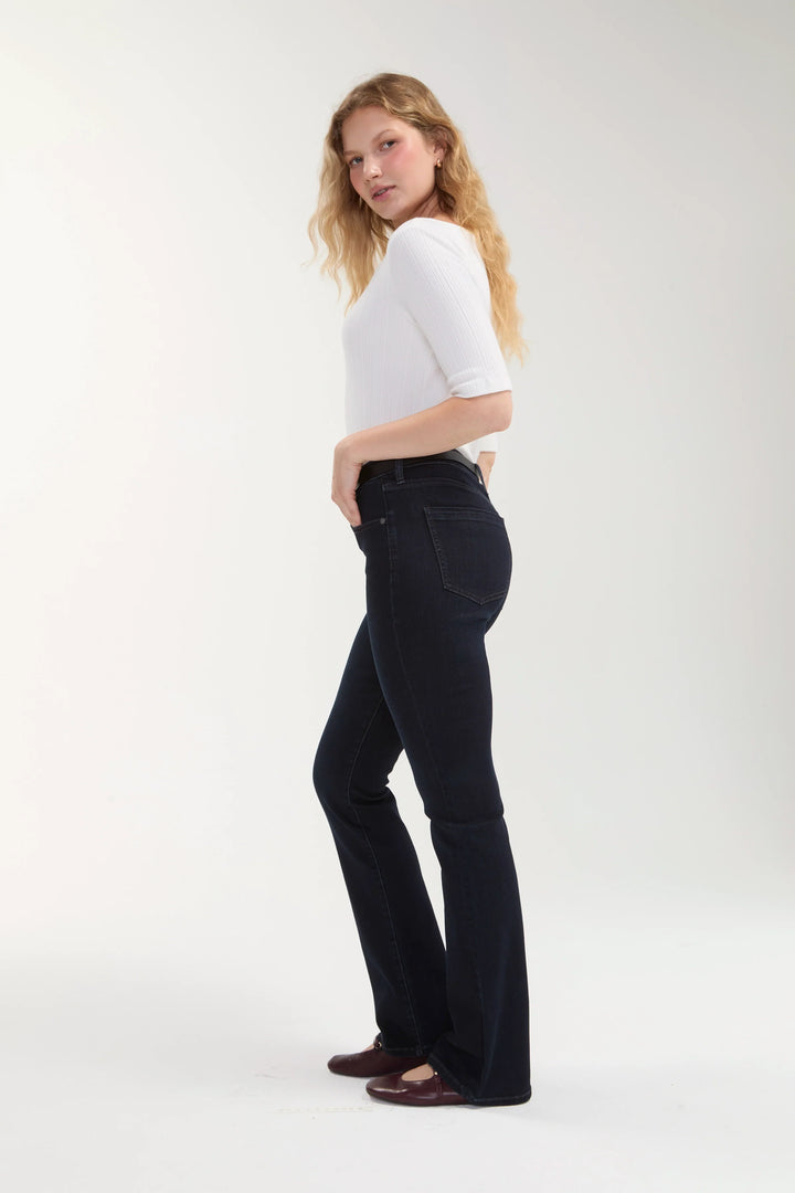 A woman modeling dark wash flared jeans with a white short-sleeve top, standing in profile against a light gray studio background.