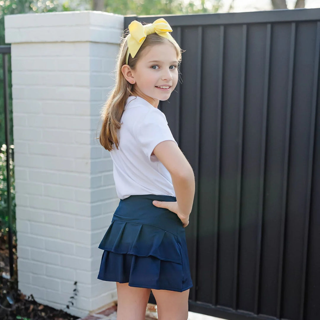 Young girl in a white shirt and navy skirt with a yellow bow standing against a white brick wall.