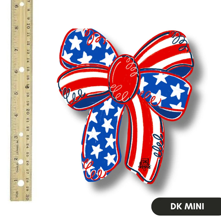 A patriotic-themed mini bow decoration with a red, white, and blue American flag pattern, shown next to a wooden ruler for scale on a white background.