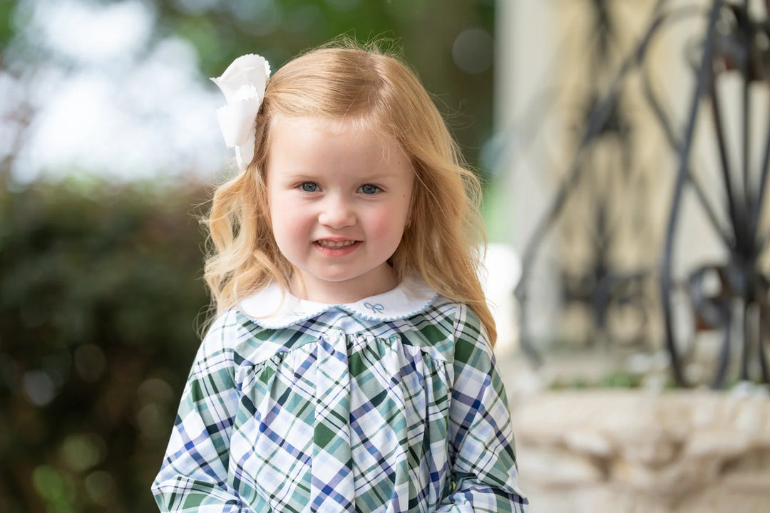 Young girl with a white bow in her hair, wearing a green plaid dress, standing outdoors.