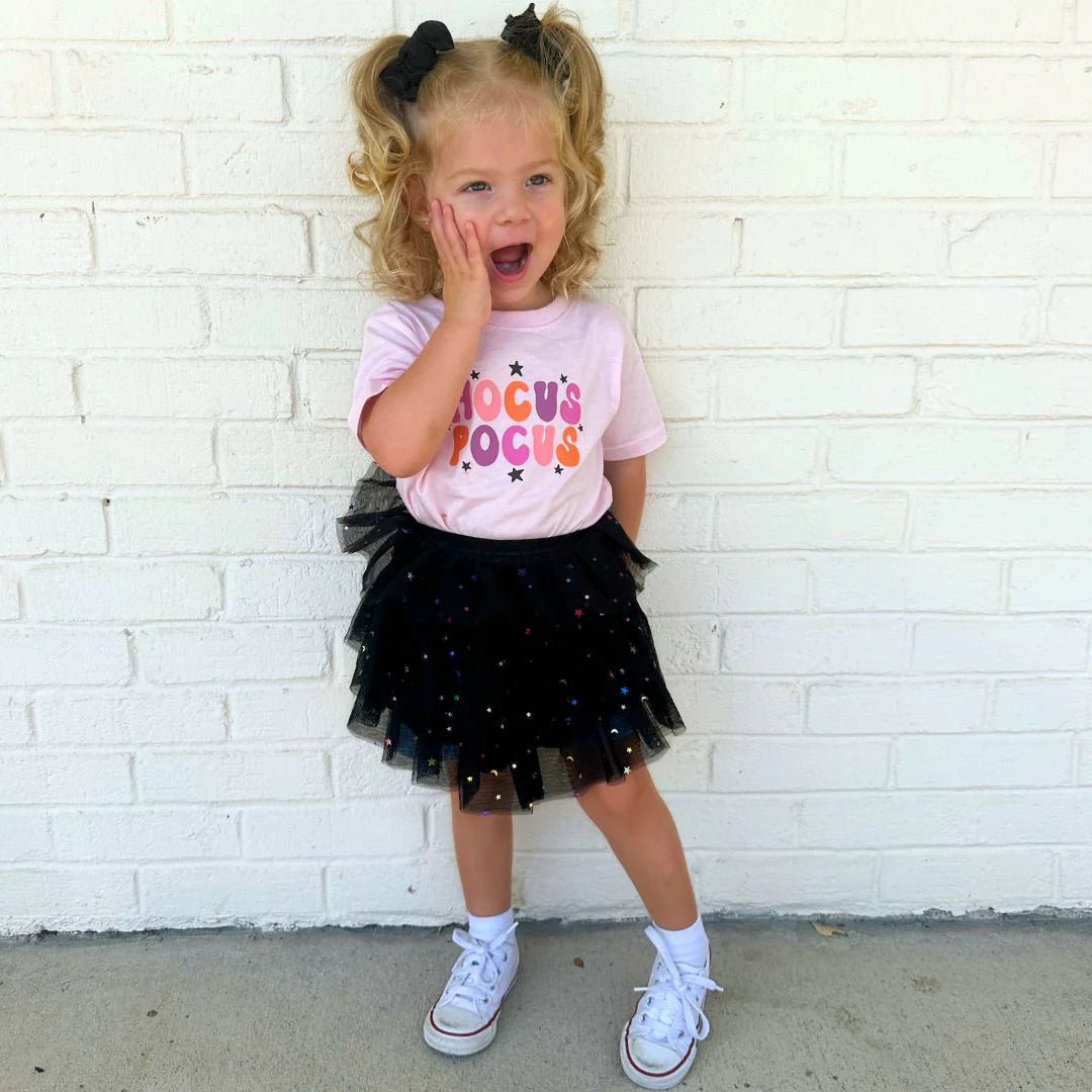 Child wearing a pink shirt with colorful text and a black tutu skirt against a white brick wall.