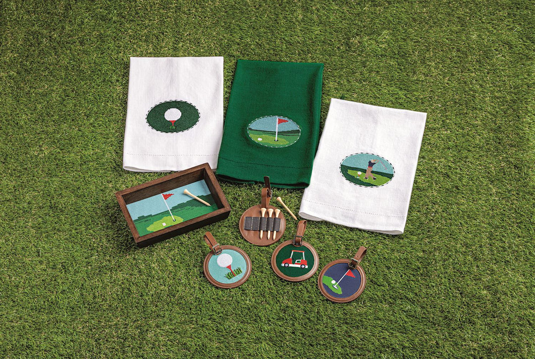 NEEDLEPOINT GOLF TOWELS, TWO WHITE AND ONE GREEN ON GREEN GRASS WITH OTHER GOLF ITEMS LAID AROUND THEM