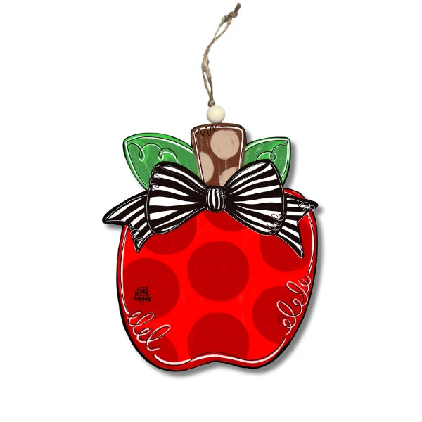 A decorative apple-shaped ornament with a glossy red body, dark red polka dots, green leaves, a black-and-white striped bow, and a hanging string on a plain white background.