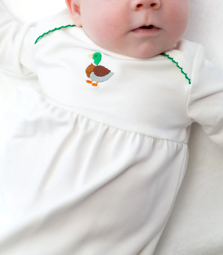 Baby wearing a white gown with an embroidered duck design on a plain background
