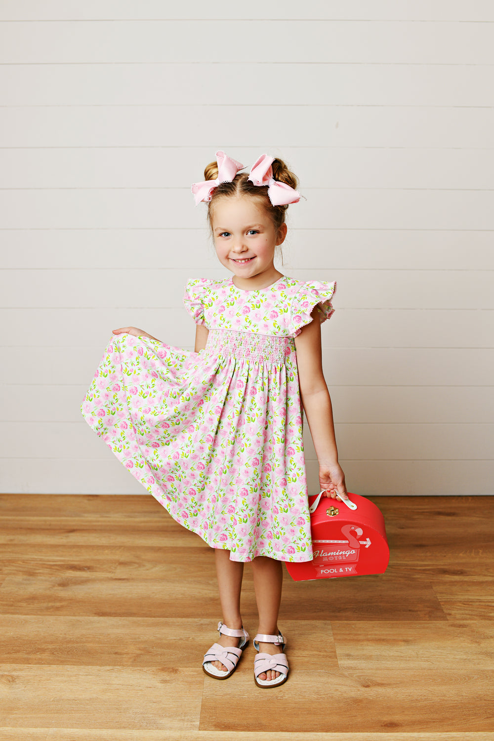 Young girl in a floral dress holding a red bag on a wooden floor.