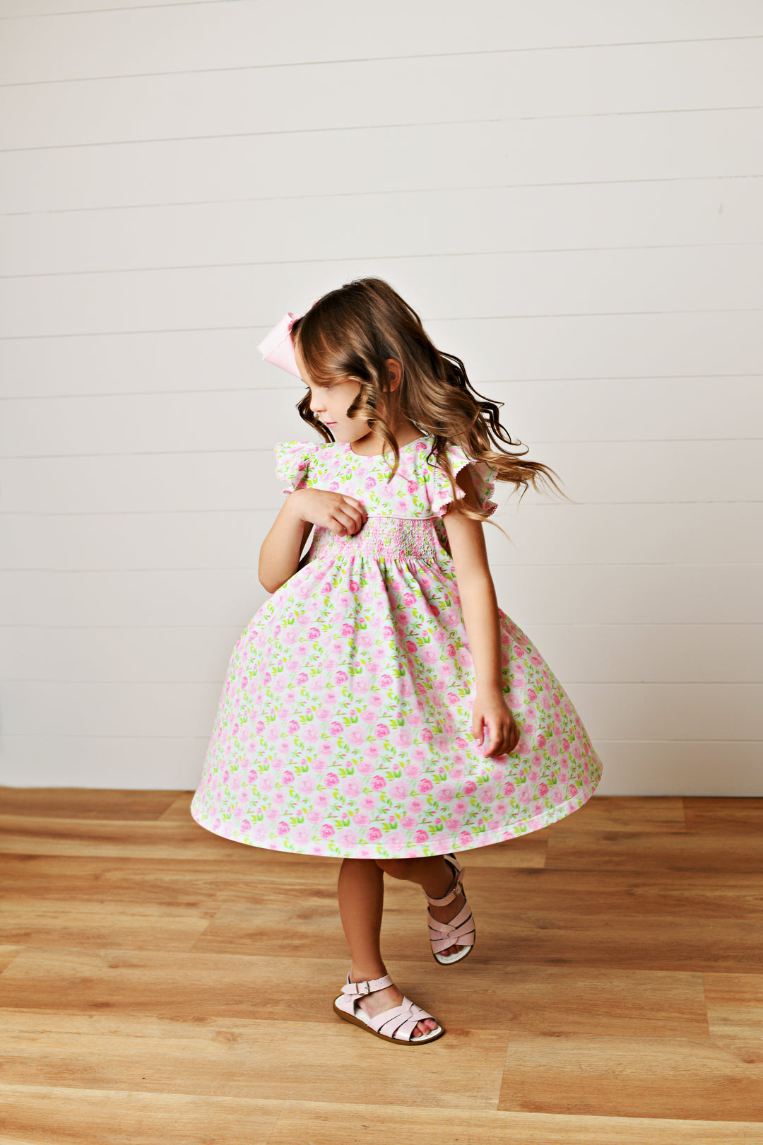 Young girl in a floral dress standing on a wooden floor with a white wall background