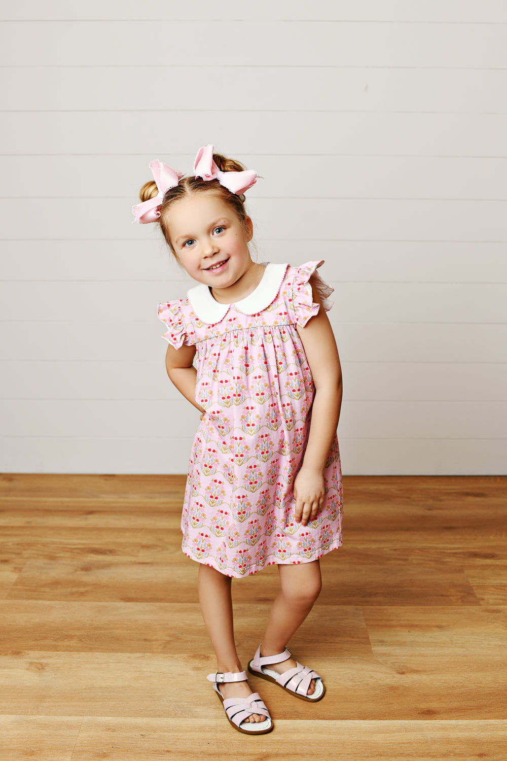 Young girl in a pink floral dress standing on a wooden floor with a white wall background