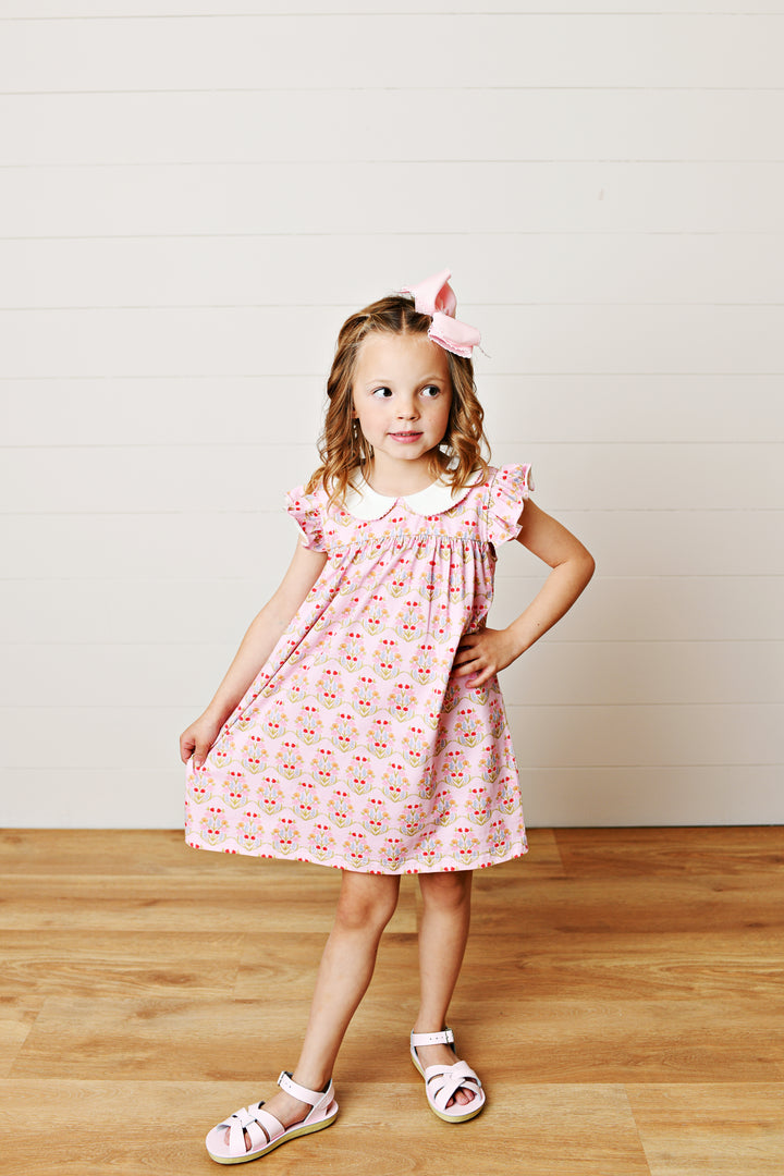 Young girl wearing a pink floral dress standing on a wooden floor with a white wall background
