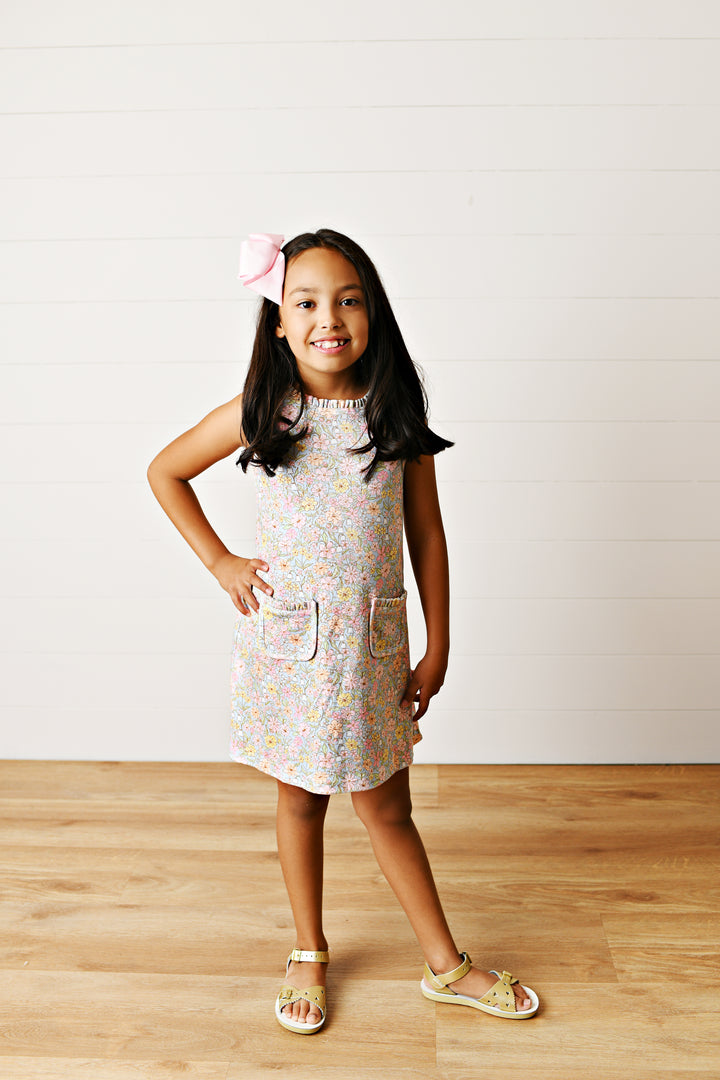 Young girl wearing a floral dress standing on a wooden floor with a white wall background