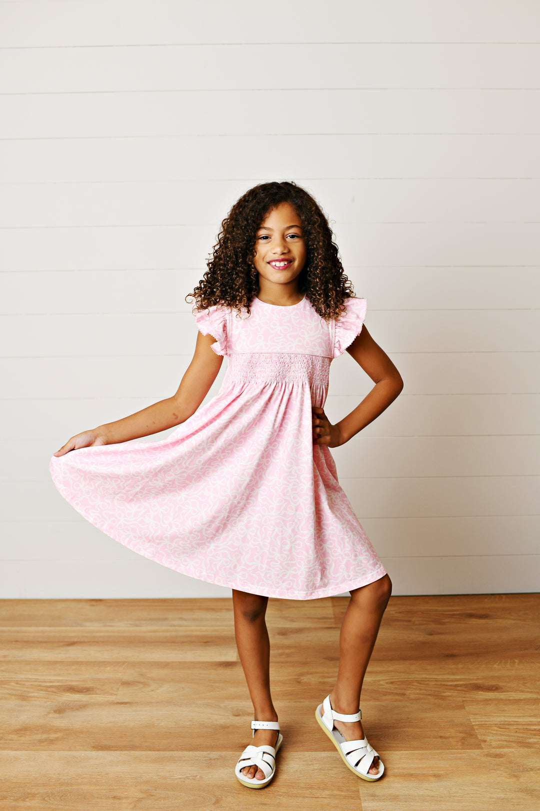 Young girl wearing a pink dress standing on a wooden floor with a white wall background