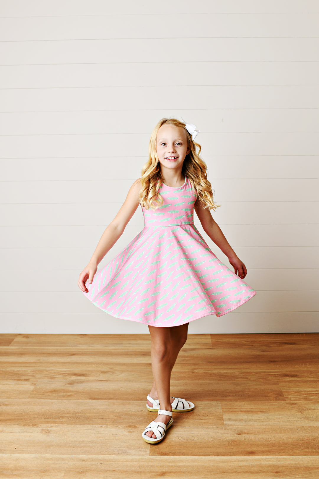 Young girl in a pink dress with white polka dots standing on a wooden floor against a white wall.