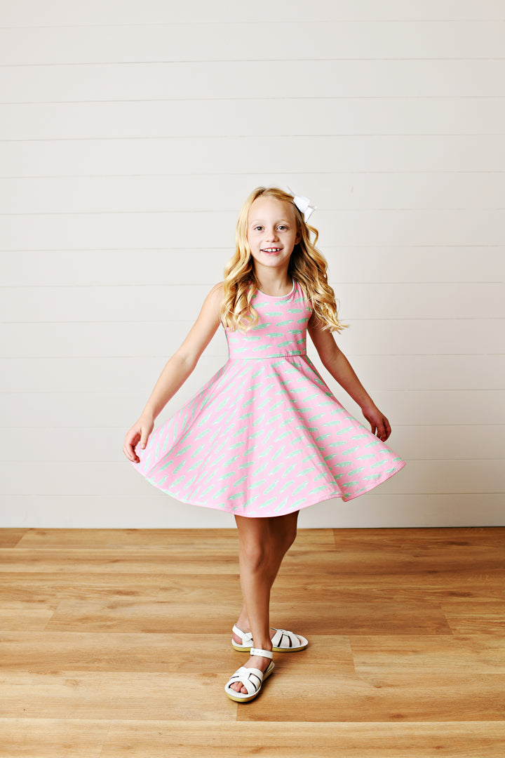 Young girl in a pink dress with white polka dots standing on a wooden floor against a white wall.