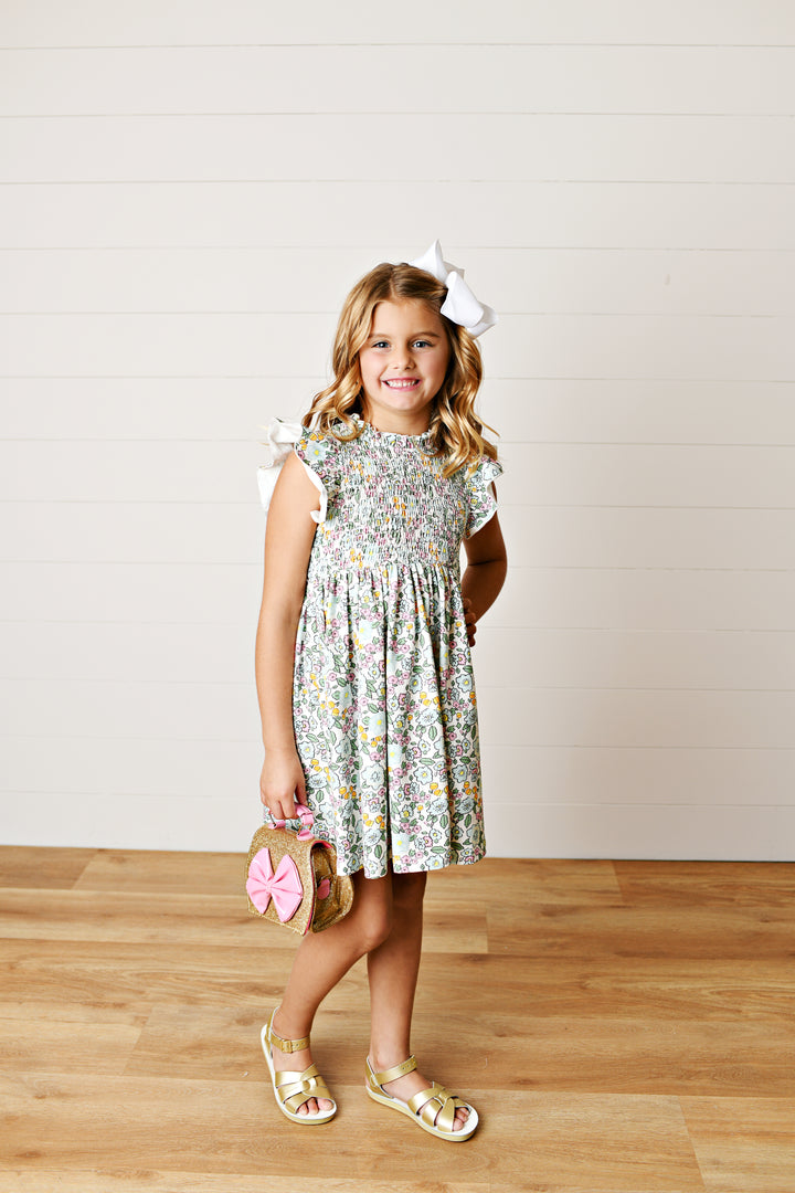 Young girl in a floral dress holding a pink bag on a wooden floor.
