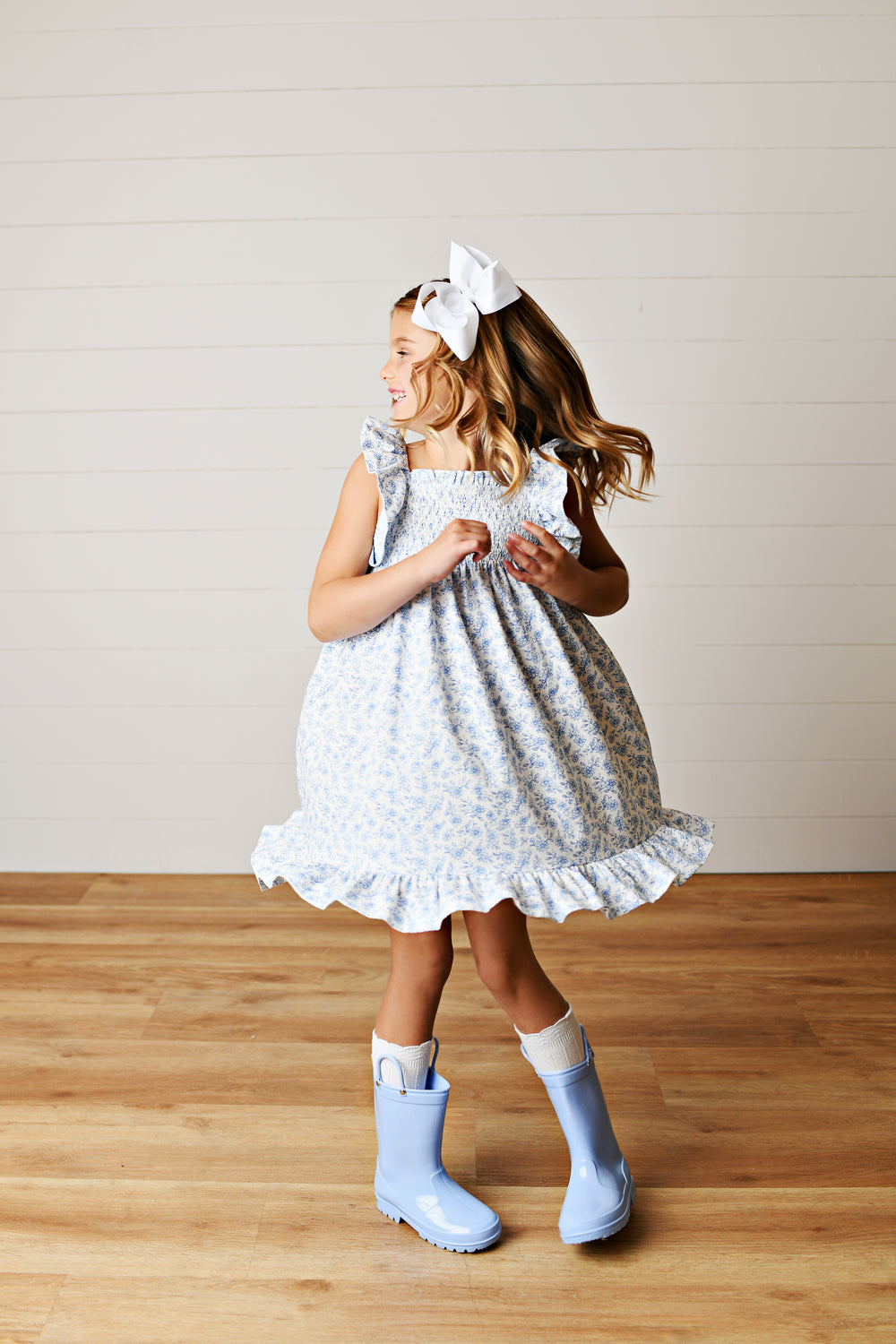 Young girl in a light blue dress with white ruffles standing on a wooden floor.