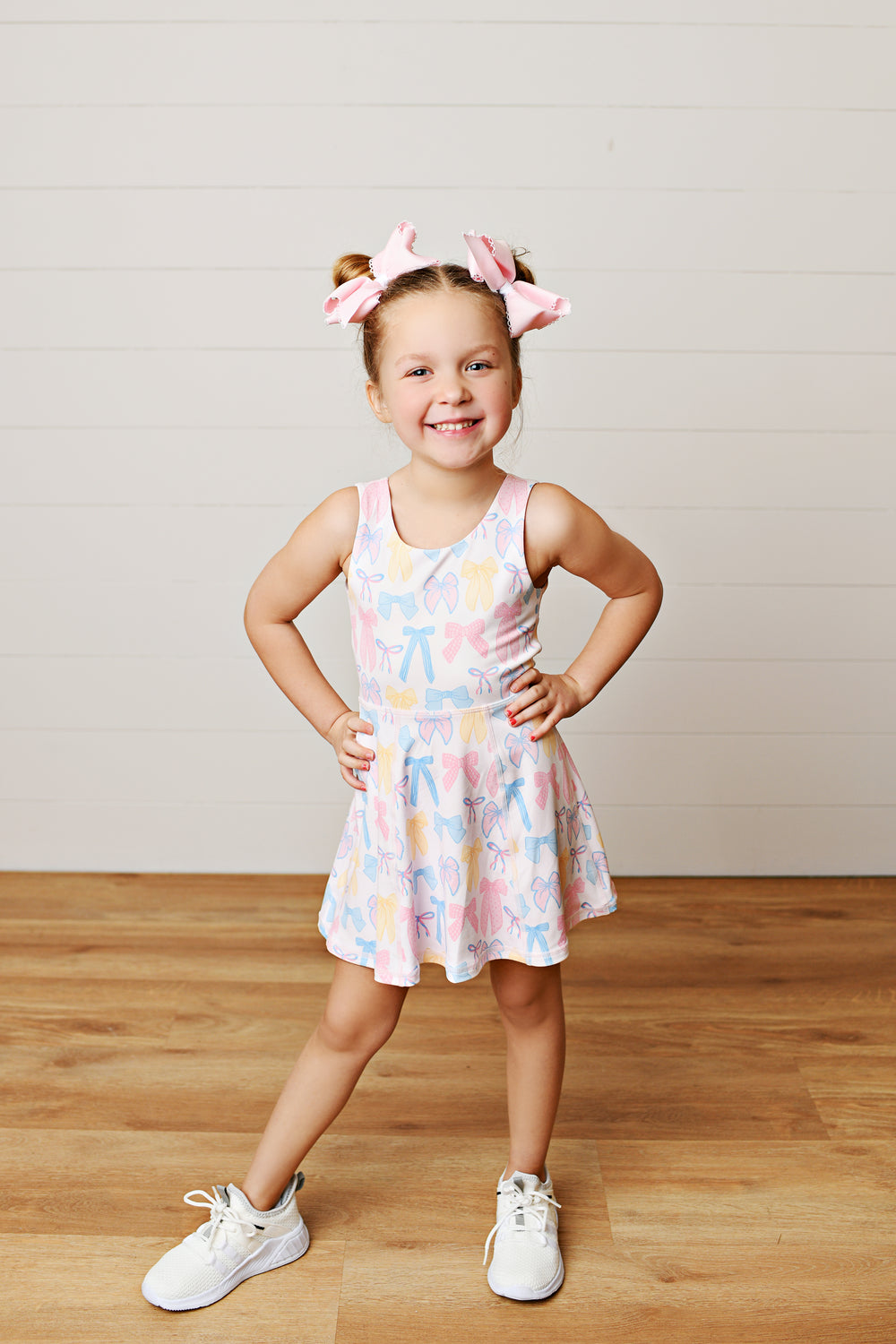 Young girl wearing a patterned dress standing on a wooden floor with a plain background
