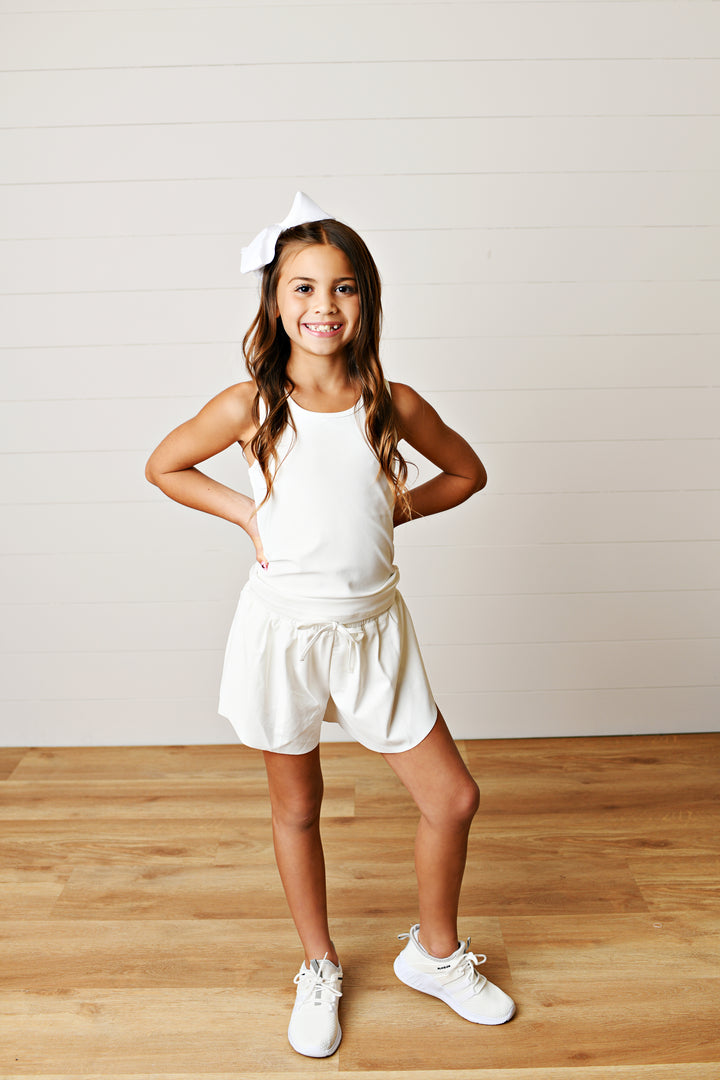 Young girl in a white outfit with a headband, standing on a wooden floor against a plain wall.