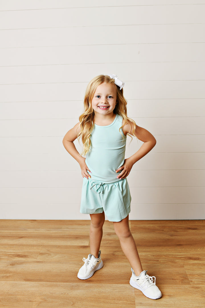 Young girl in a light green outfit standing on a wooden floor with a white wall background