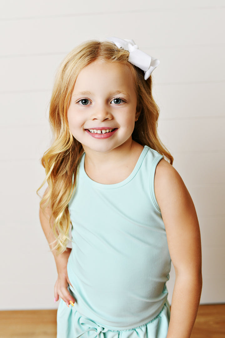 Young girl wearing a light blue dress with a white bow in her hair against a plain background