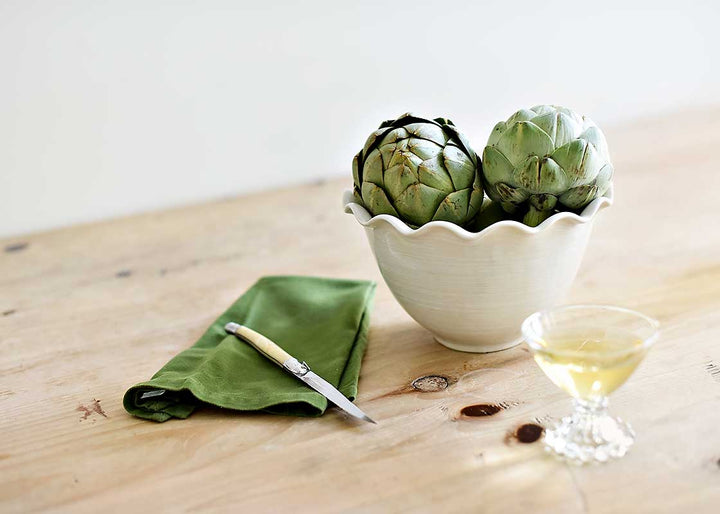 Two artichokes in a white bowl on a wooden table with a green napkin and knife.