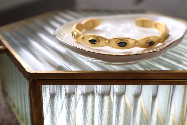 A gold cuff bracelet with oval links and dark oval gemstones, displayed on a white ceramic dish resting on a ribbed glass table with a gold frame.