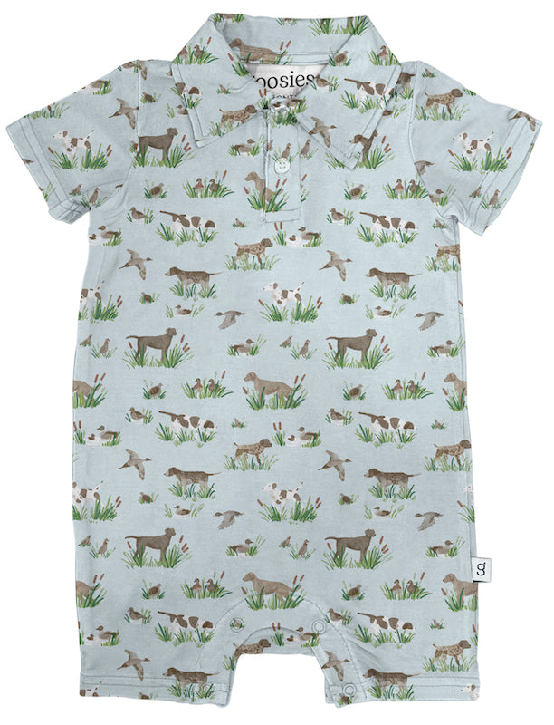 A baby short-sleeve romper with a light blue-gray background and an all-over print of dogs in fields with green grass and brown flowers. The brand name 'nosies' is visible on the inner neck label.