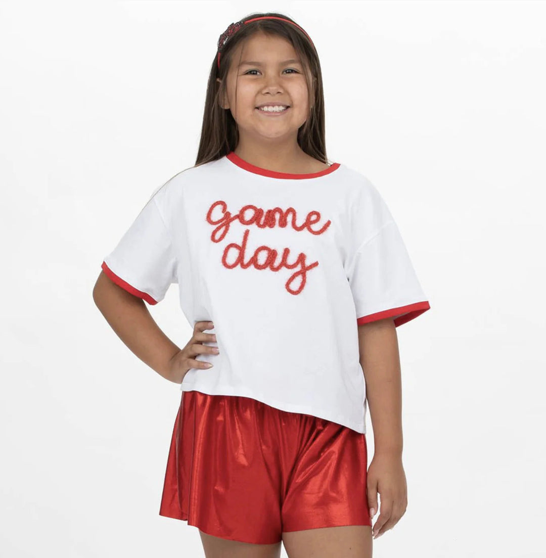 Young girl wearing a 'game day' shirt and red shorts on a white background