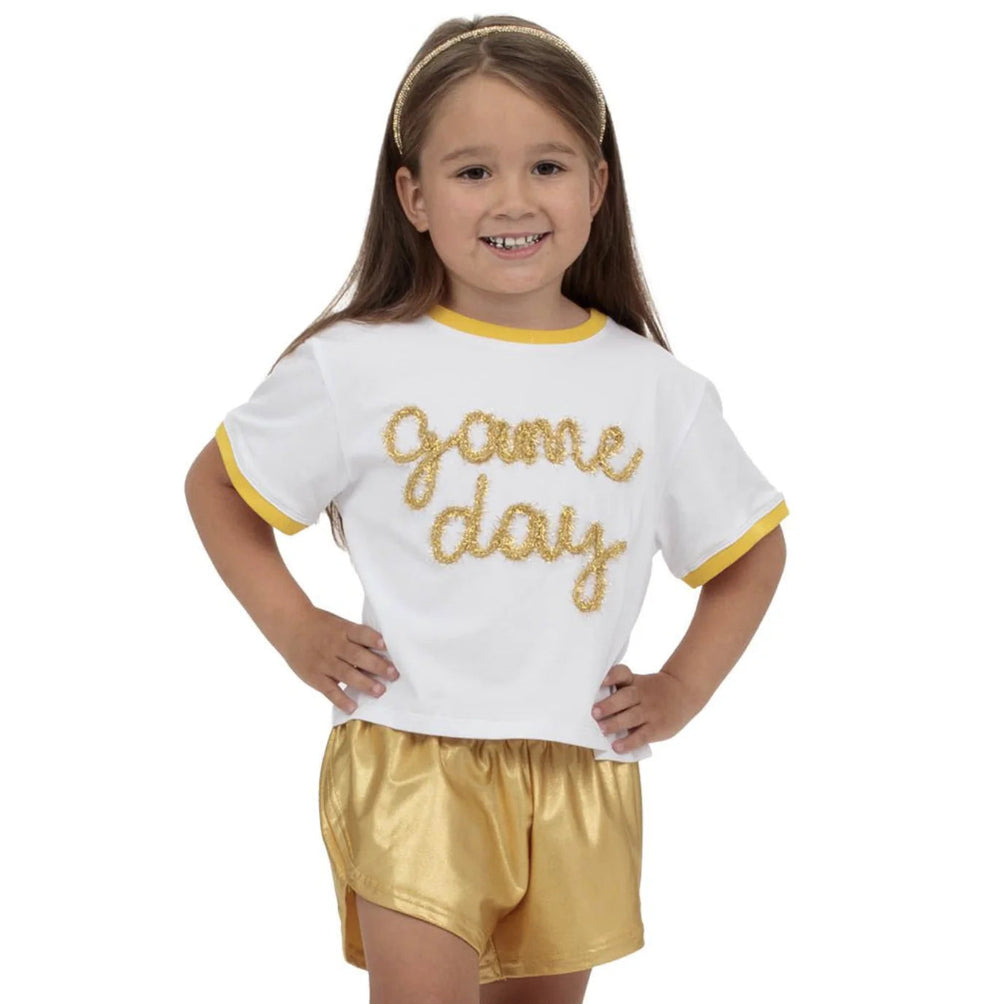 Young girl wearing a 'game day' outfit with a white shirt and gold shorts on a white background.