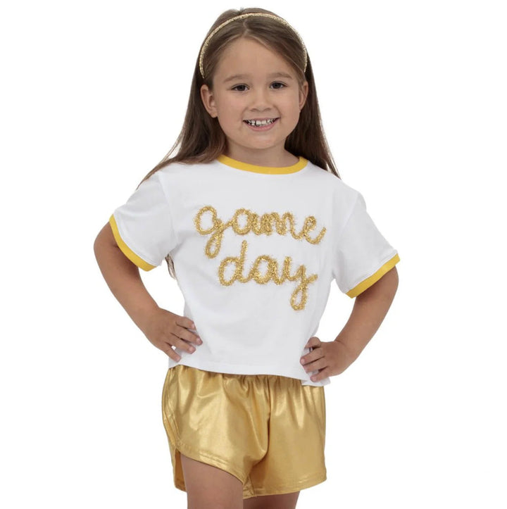 Young girl wearing a 'game day' outfit with a white shirt and gold shorts on a white background.