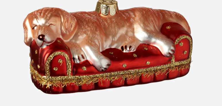 Decorative glass ornament of a dog lying on a red couch with gold accents.
