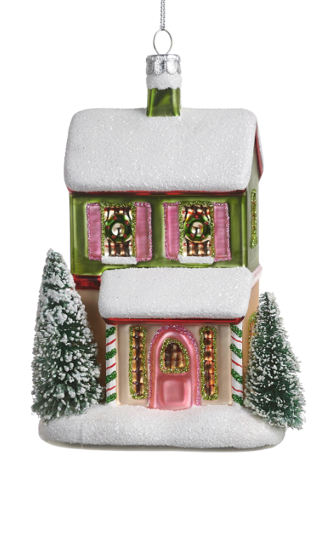 A Christmas house ornament with a snow-covered roof and base, pink shutters and door, green trim, candy cane details, and two small frosted evergreen trees on a white background.