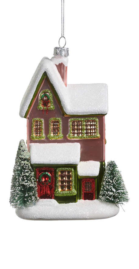 A Christmas house ornament with a brown body, white snow-dusted roof and ground, green trim around the windows and wreath, and red doors and window accents, hanging against a white background.