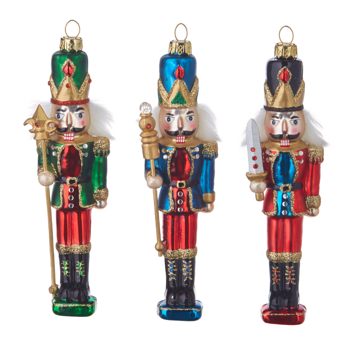 Three traditional Christmas nutcracker ornaments standing side by side on a white background. The left nutcracker wears green and red with gold accents and holds a spear.