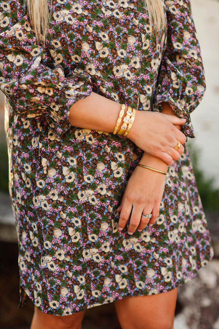A close-up of a woman wearing a long-sleeve floral dress with a dense print of small white, purple, and yellow flowers on a dark green background. She is accessorized with gold bracelets and rings, and her hands are crossed over her waist.